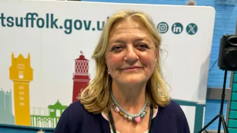 A woman is standing in front of a county council poster and speaker. She is smiling at the camera and is wearing a blue cardigan and a green and pink necklace. 