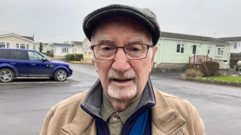 Dennis Elliott is standing in a car park in front of residential houses. He is wearing a brown jacket, glasses and a hat.