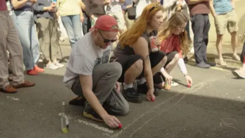 Oliver Dupont Three people kneel down behind a white line holding red Babybels, ready to roll them down the hill, with spectators stood around.