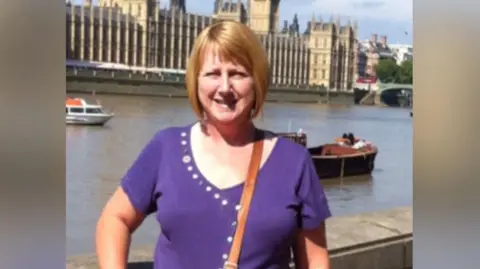 Family handout Susan Spring looks at the camera wearing a purple v-neck, short sleeved top. She has a brown handbag crossed over her shoulders. She looks at camera with strawberry blonde bobbed hair which she wears in a bob with a fringe. She is smiling and is standing in front of the Thames opposite the house of parliament