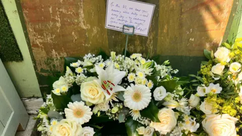 A floral wreath made up of light yellow and white flowers with a message attached saying: "To the 72. We keep your memory alive today and every day by continuing our campaign for answers, justice and change. Mayor of London"