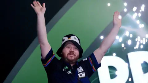 Justin Hood celebrates with his hands in the air at the World Darts Championship. He is wearing a black bucket hat with a cartoon penguin print.