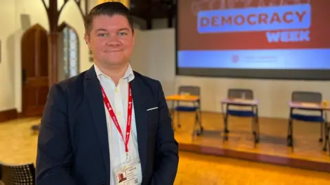 Sam smiles at the camera with a projector with Democracy Week projected on it. The stage with the seats and tables politicans were using are in the background. Sam is wearing a dark blue jacket with a white shirt and he has a visitor lanyard on with a red collar.