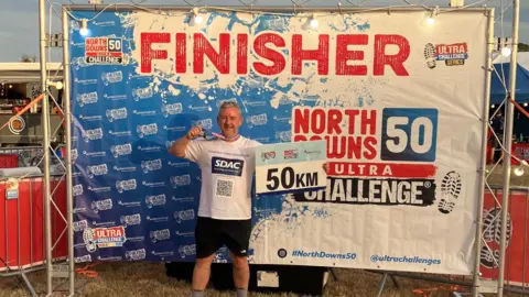 David Kerr A man in white T-shirt and black shorts, holds a medal aloft with a 50km sign in front of a Finisher sign for the the North Downs Ultra Challenge