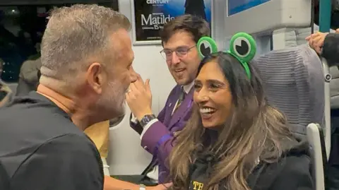 LNR Steve and Payal, both smiling, with a man with a purple jacket, clapping, looking at them. They are all sitting on a train with other passengers on the train. 
