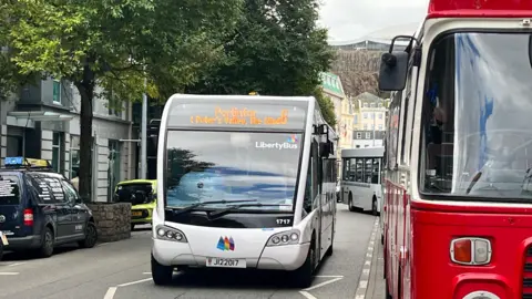 BBC A small white bus travelling through a street. A red bus is parked at a bus stop on the right-hand side. Cars are parked on the left hand-side. Trees line the streets. Buildings can be seen in the background.