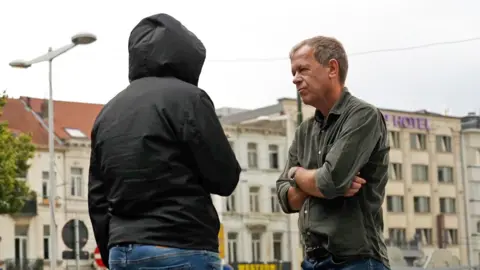 BBC Man in black coat with hood up, with his back to us stands opposite Andrew Harding who is wearing a green shirt and has his arms crossed.