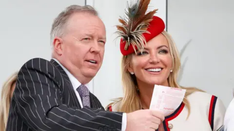 Getty Images Doug Barrowman, a man with greying hair, is wearing a grey chalk-stripe suit and white tie with purple geometric pattered shirt. He is holding a number of betting slips. Michelle Mone is in a cream, red and black jacket with matching hat. She has blonde hair and is smiling.