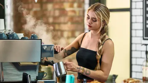 A barista holding a silver metal milk jug against a coffee machine. She has blonde hair in a plait and it wearing a black vest top. She also has tattoos on both arms