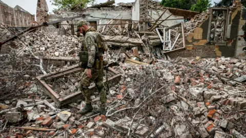 Shutterstock A Ukrainian soldier stands on large piles of rubble where a house once stood. There are bombed buildings in the background