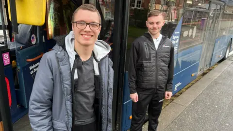 BBC/RICHARD EDWARDS Tyler and Alfie stand by the side of a double decker bus with its doors open. Tyler is wearing glasses and a grey coat. Alfie is wearing a black hoodie and a white T-shirt. 