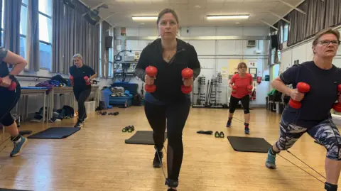 BBC A woman in a black gym top and black leggings holding two red dumbbells