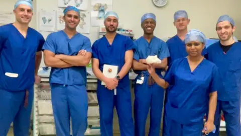 University Hospitals Birmingham NHS Foundation Trust Seven people are wearing blue healthcare uniforms and looking at the camera. There are six men, near a wall that includes a clock, and a woman is in front of them.