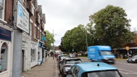 Christopher Day/LDRS Harpenden High Street with a row of cars parked on the left and a blue van driving past on the other side, trees in the background