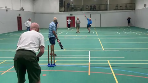 Gloucestershire Cricket Foundation A group of older men play cricket in a sports hall, a man is hurling a ball towards a batter. They are dressed in plain clothing as it is walking cricket.