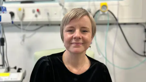 A woman with short blonde hair, ear rings and a necklace and a black top is sitting in a hospital environment. There are an assortment of different coloured tubes and a technical machine out of focus behind her.
