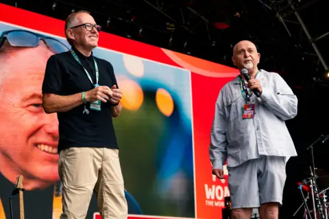 Two men stood smiling on a stage with lanyards on which say 'Womad'