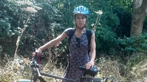 Louise Weissel A woman - Louise Weissel - standing and holding her bike wearing a blue bike helmet and a navy blue printed dress, looking straight-faced, with trees and hedgerows in the background. 