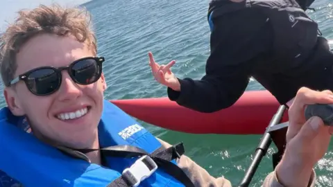 Alfie Watts Alfie is smiling, wearing sunglasses and wearing a blue life jacket - he is on the water. His left hand can be seen holding a handle, and you can see there is someone behind him in black, with a red long float keeping them afloat. It is a sunny day.