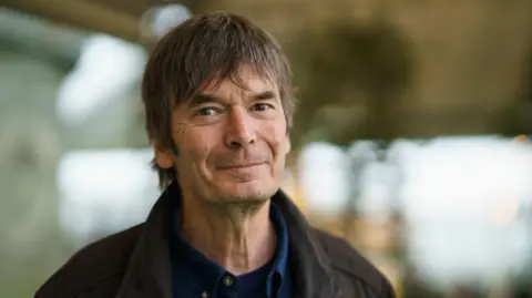 Getty Images Ian Rankin. He has brown hair and dark eyes and is wearing a brown jacket and blue shirt. The background is blurred.