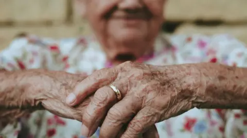 A smiling person wearing a ring holds their hands together in a relaxed manner