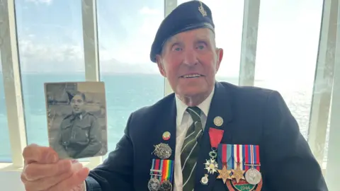 Olivia Richwald/BBC A old man wearing military medals smiles as he holds a black and white photo of a young man in uniform. The sea is in the background.
