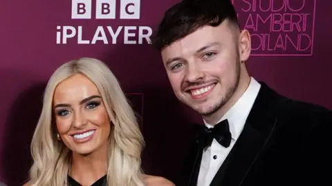 PA Project manager Jake Brown and former soldier Leanne Quigley in formal attire smiling at a photocall at the BBC. Jake is wearing a black bow tie.