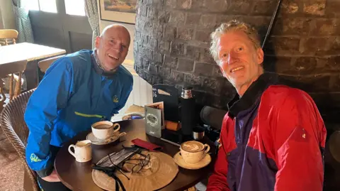 Two men smile for the camera while sitting around a table. They both have a coffee in front of them. ?They are dressed in waterproof clothing.