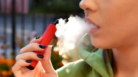 A woman with black nail polish holding a red vape exhaling smoke. She is wearing a green hoodie.