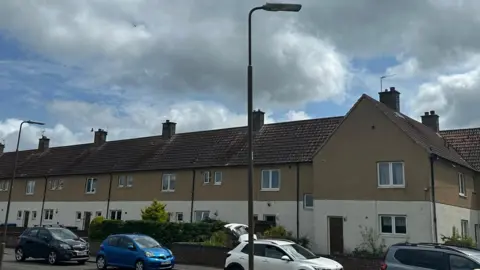 A row of one-storey houses, cream on the bottom floor, brown on the top, with cars parked in the road outside.