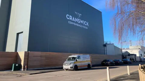 The outside of the factory. The words "Cranswick Convenience Foods" are written in white lettering on the side of the building. An "abnormal load" van and a few cars are parked outside