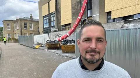 Kent County Councillor James DeFriend stands in front of the fire ravaged Conqueror House on Crabble Hill, Dover. A rubble chute, temporary fencing, boarded up windows and smoke damage cover the apartment building