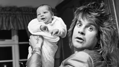 Getty Images A black and white image of Ozzy looking startled, holding up a young baby dressed in white. The baby's face is crinkled. Ozzy has long layered hair and appears to be wearing a silk suit. A window valance can be seen behind.
