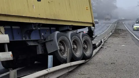 A lorry's rear wheels crushing part of the central reservation on the A14. There is a yellow container on the lorry. Cones can be seen cordoning off a lane on each side of the road. 