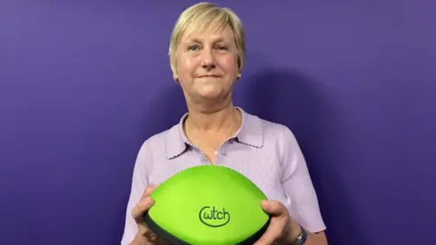 A woman with short blonde hair stands in front of a purple background wearing a lilac tshirt. She's holding a neon green oval case with the word Cwtch on it.