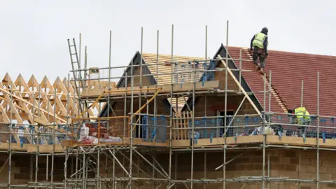 Two workers wearing hi-vis jackets work on the roof of a new, partially built house on a construction site.