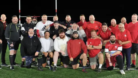 Man v Fat Rugby Cheltenham A group of about 20 men posing for a photograph together on a flood-lit rugby pitch at night, smiling to camera, some in white kit and some in red, with others i black sports gear
