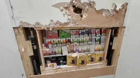 Essex Police A large collection of illegal vapes in colourful boxes stashed behind a wall, which has been peeled back by officers.