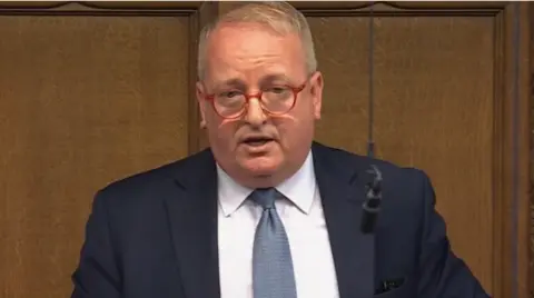 House of Commons A man with short fair hair and red-framed glasses stands in the House of Commons in a navy blue suit, white shirt and blue tie. The wood panelling of the chamber is behind him, and a microphone hangs on a wire in front of him.