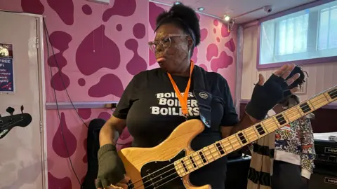 Christine stands in the rehearsal studio holding her guitar 
