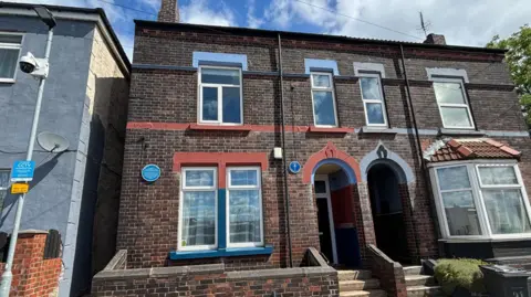 Simon Thake/BBC The exterior of a dark semi-detached brick house with blue and red painted edges