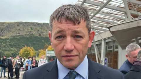 David Threadgold lasting  extracurricular  the Scottish Parliament building. He is looking consecutive  into the camera. He is wearing a suit, with a airy  bluish  garment  and acheronian  tie. 