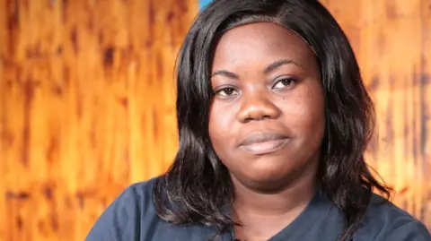 A woman wearing a navy blue T-shirt sits in a wood-panelled room.