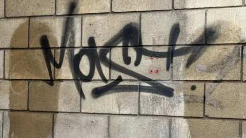 A photo of tagging graffiti on a wall in Cheltenham. The tag has been spray painted using black paint and the wall is a mixture of beige-coloured brick and white