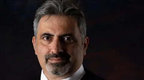 Antony Antoniou Antony Antoniou with dark hair and beard against a dark background