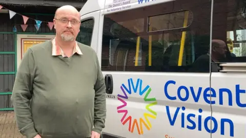 A bearded man in a green jumper and glasses stands outside, next to a 'Coventry Vision Hub' minibus.
