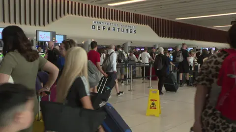 A crowded departure lounge at Manchester Airport with passengers pulling their luggage along and queueing at check in