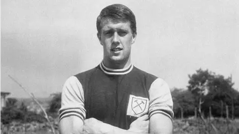 Getty Images Sir Geoff Hurst, who has his arms folded, is staring at the camera in in his West Ham kit in 1960. 