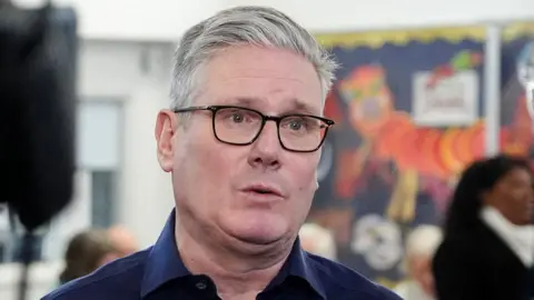 Reuters Sir Keir Starmer wearing black glasses and a blue shirt as he speaks to a person out of frame with a camera to his right and the classroom in the background as he visits Emmer Green Youth & Community Centre in Reading on Monday.