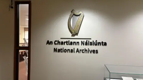 National Archives of Ireland sign at their archive offices in Dublin. A decoration of a gold harp is on the wall. 
